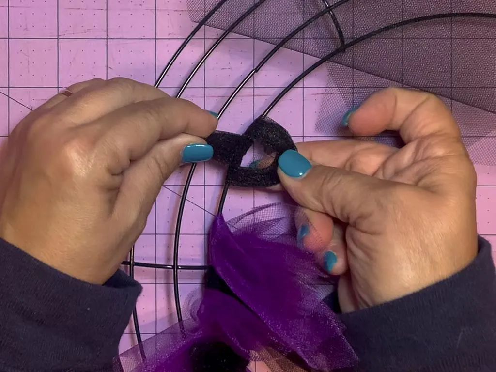 Two hands pulling the tail of tulle through the loop in the process of tying a tulle slip knot on a wreath form.