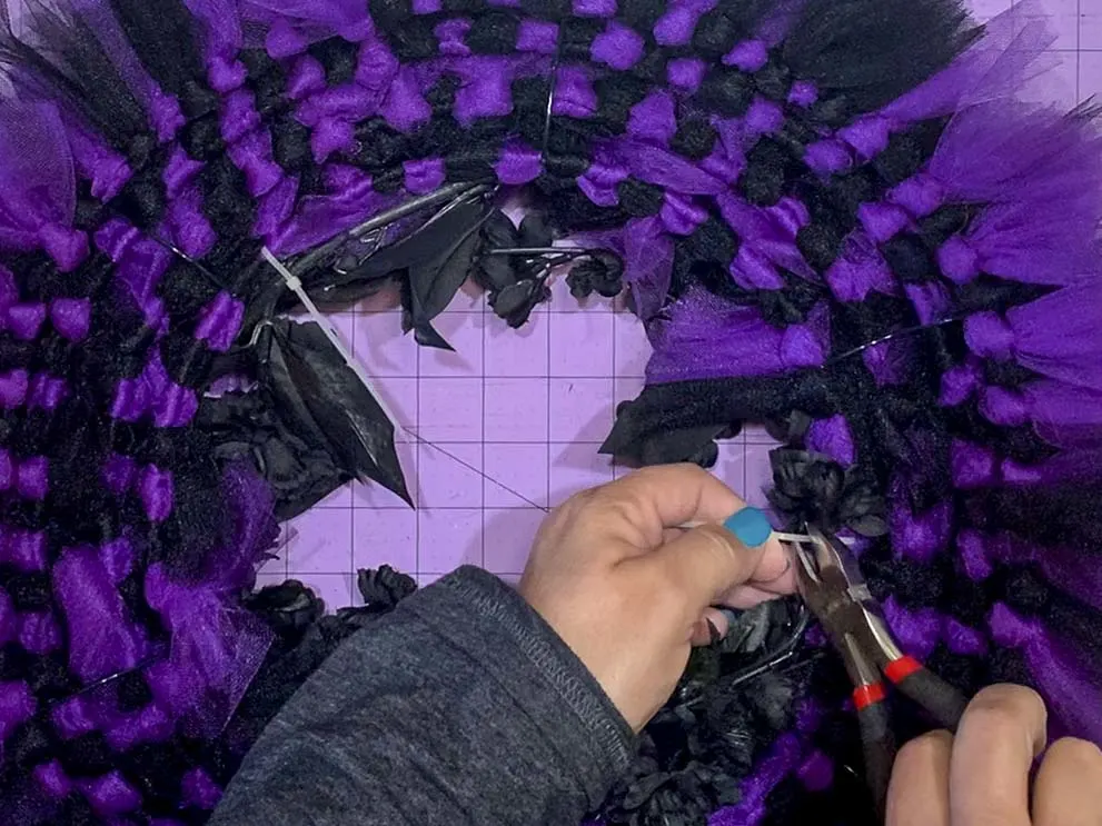 Hands using wire cutters to snip off the excess piece of a zip tie used to attach an artificial flower to the back of a tulle wreath.