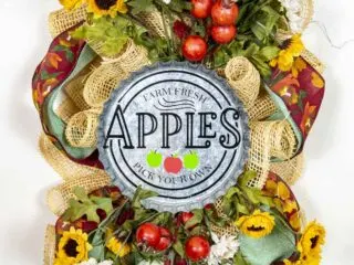 A fall swag made of poly burlap mesh decorated with ribbons, apples and flowers in autumn colors. Features a round metal sign saying “Farm Fresh Apples – Pick Your Own.”
