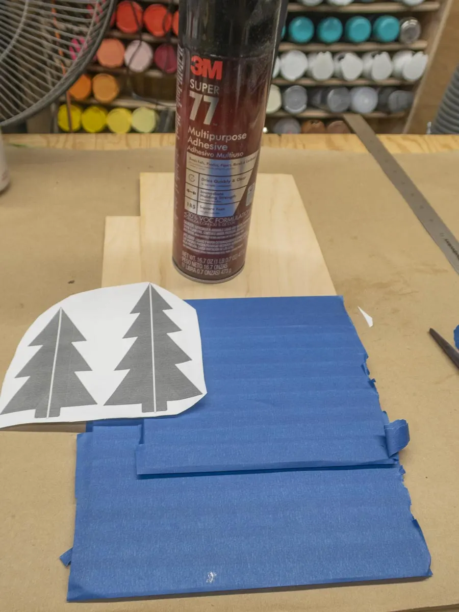 Template for cutting 3D pine trees, two pieces of wood covered in painters tape, and a can of 3M 77 spray adhesive for attaching the template to the taped wood.