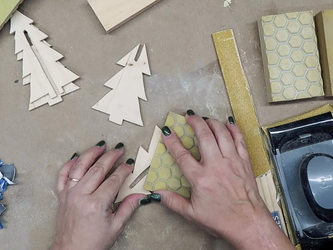 Hands using a sanding sponge to sand the wood tree cutouts.