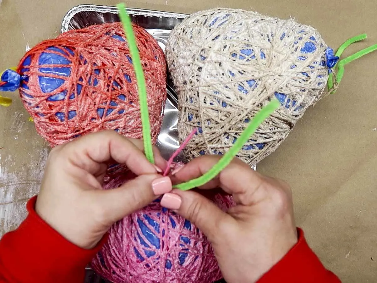 Hands tying a pipe cleaner to the lip of the balloon to facilitate hanging the glue-covered hearts for drying.