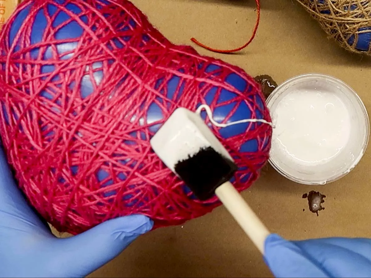 Hand applying glue to a twine-covered balloon using a foam brush.