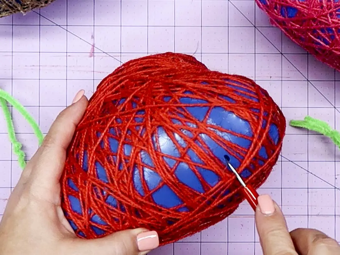 Hands using a sharp point to poke a hole in the balloon wrapped with twine after the glue mixed dried.